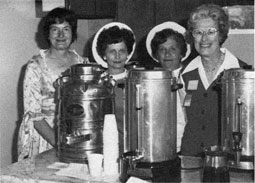 Four women with smiles standing behind 3 large coffee urns.