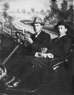 Two well dress young men in an open car with a road backdrop behind them.