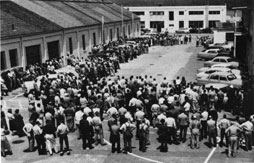A large gathering of workers standing before industrial buildings.